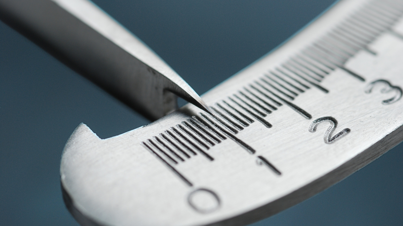 The image is a close-up photograph of a metal caliper’s jaws positioned closely around a stainless steel ruler. The caliper’s pointed tip is accurately aligned with the fine, etched markings on the ruler, indicating the number 2 in millimeters. The background is a smooth, muted blue, emphasizing the sharpness and clarity of the metallic tools in the foreground. This image visually represents the concept of precision in measurement, crucial for maintaining high standards of data accuracy in technical and scientific fields.